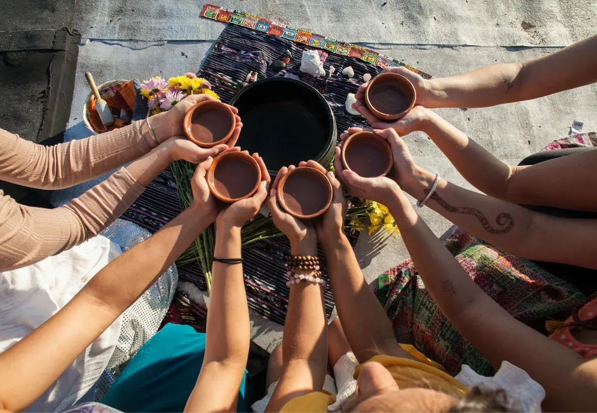 Hands holding traditional clay cups of ceremonial cacao raised together in circle over woven textiles and flowers