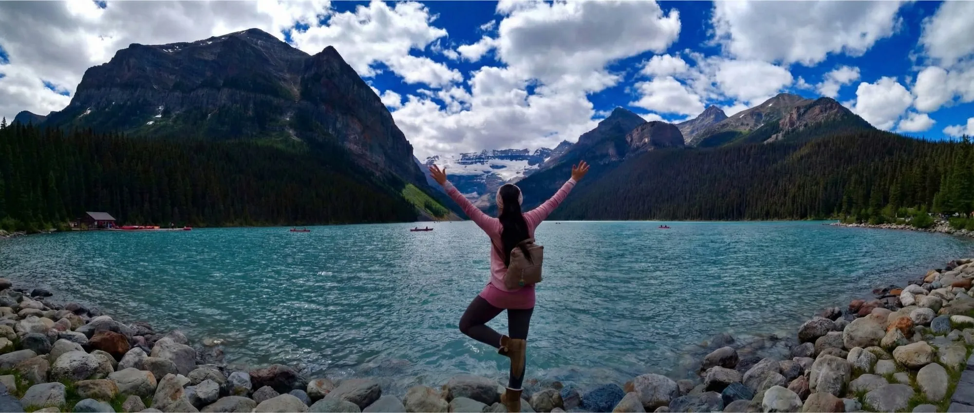 Silvia in tree pose with arms raised at a turquoise mountain lake surrounded by dramatic peaks and clouds