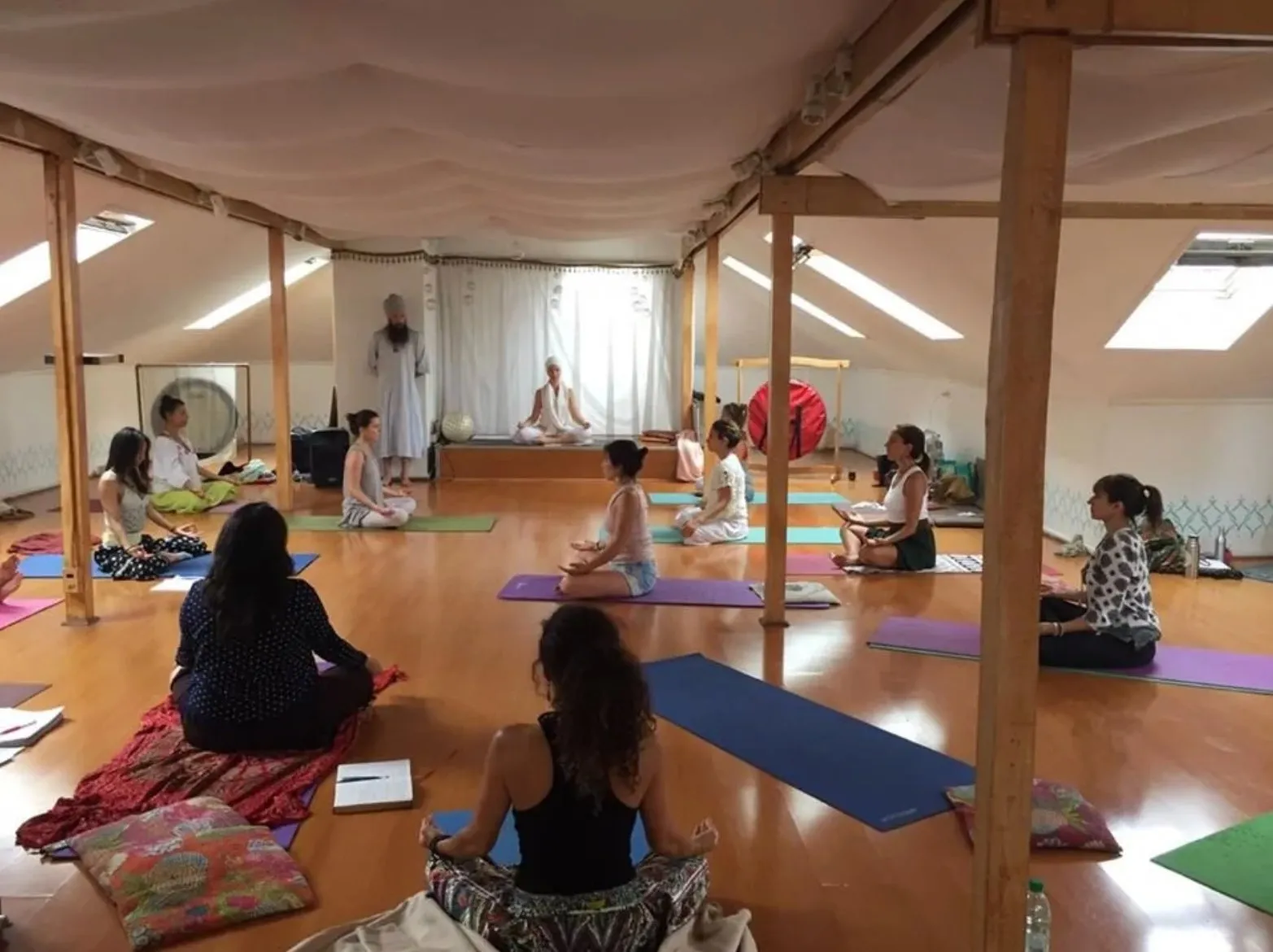 Kundalini yoga meditation class with students seated on mats in white-draped studio with gong and teachers in white